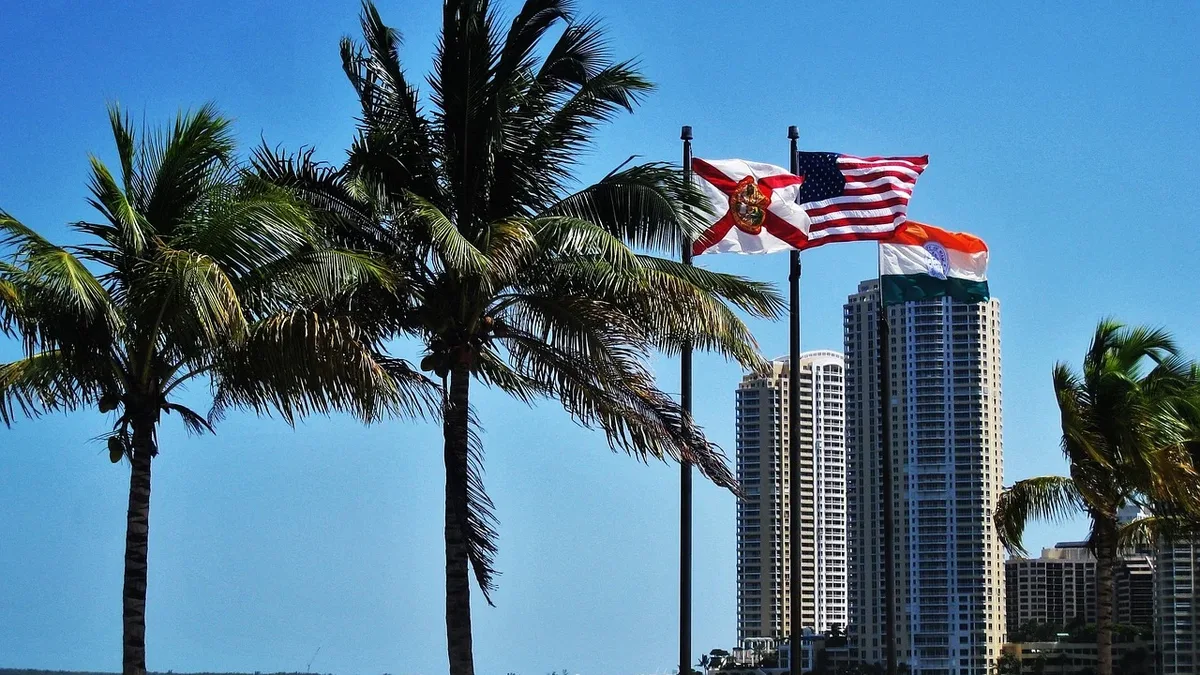 A row of palm trees stands on a beach, with two flags flying in the wind: the American flag and the Florida state flag, set against a backdrop of a large body of water and a city skyline.