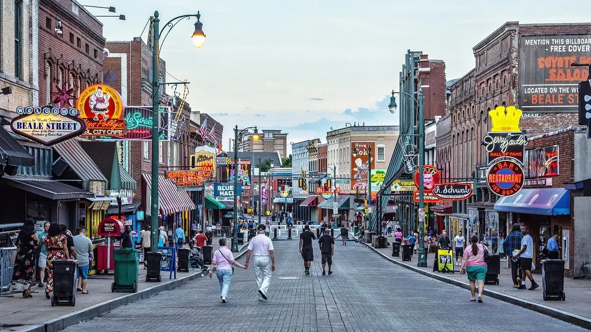 A bustling street scene in Memphis, Tennessee, featuring a wide, paved road lined with numerous shops and restaurants, with people walking along the sidewalks.